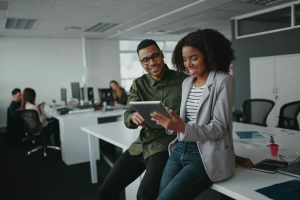Successful two african american young businesspeople sitting on desk using digital tablet while colleague in background at office Networking & Community